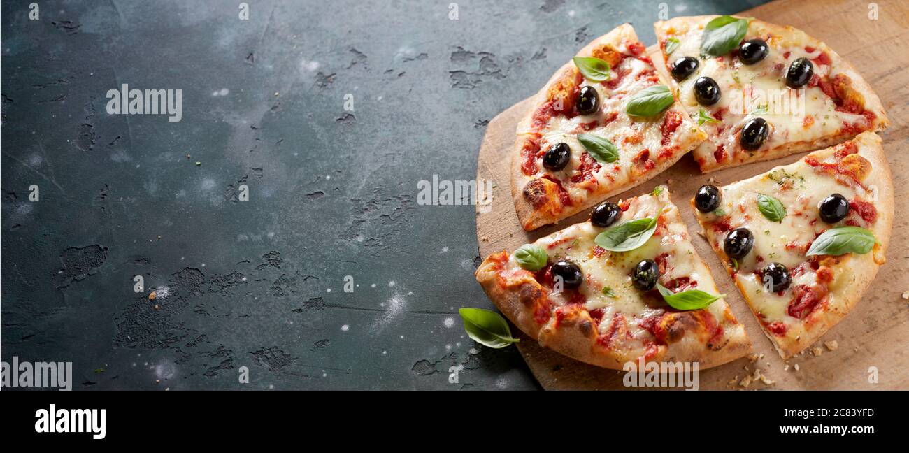 Sliced whole pizza with olives and fresh basil leaves on mozzarella cheese served on a wooden cutting board over slate in a panorama banner with copys Stock Photo