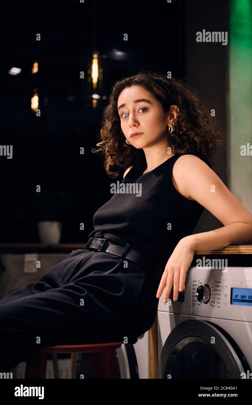 Beautiful casual girl confidently looking in camera waiting washing in self-service laundry at night Stock Photo