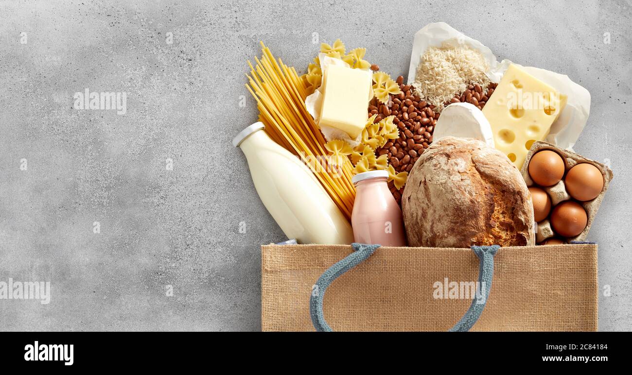 Shopping bag filled with fresh dairy products and carbohydrates such as cheese, pasta, baguette, beans, eggs and milk over a textured grey background Stock Photo