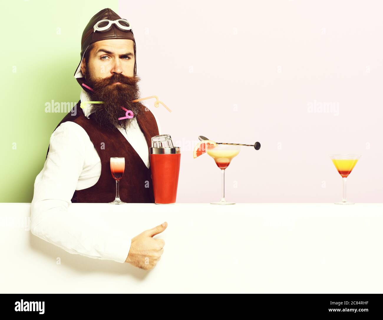 handsome bearded aviator or pilot with beard and mustache on serious face with shaker alcoholic cocktails in vintage suede leather waistcoat with hand, and glasses on purple green studio background Stock Photo