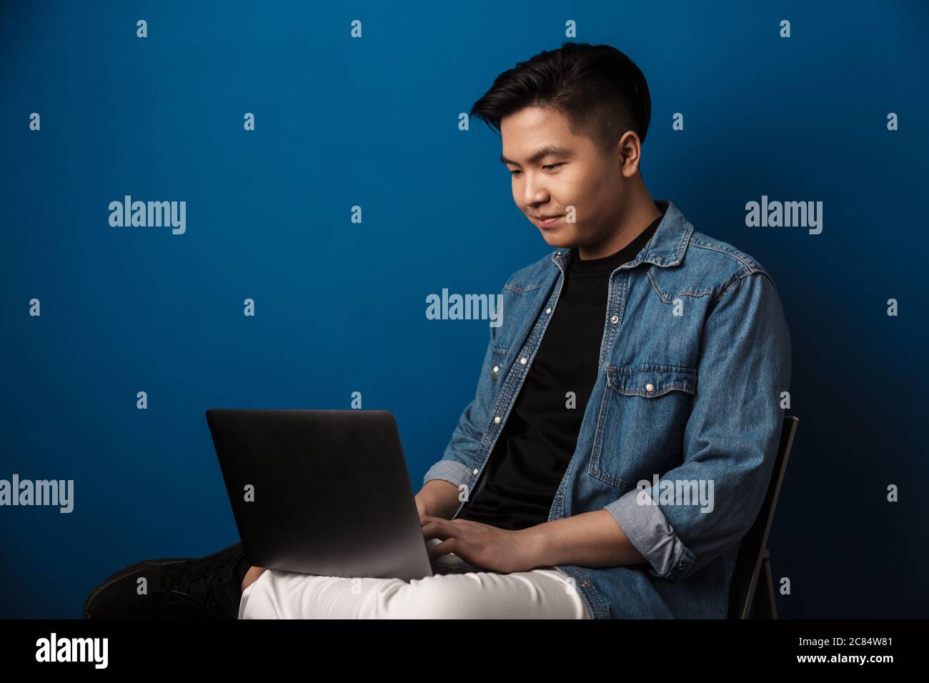 Image of pleased handsome asian man using laptop while sitting on chair ...