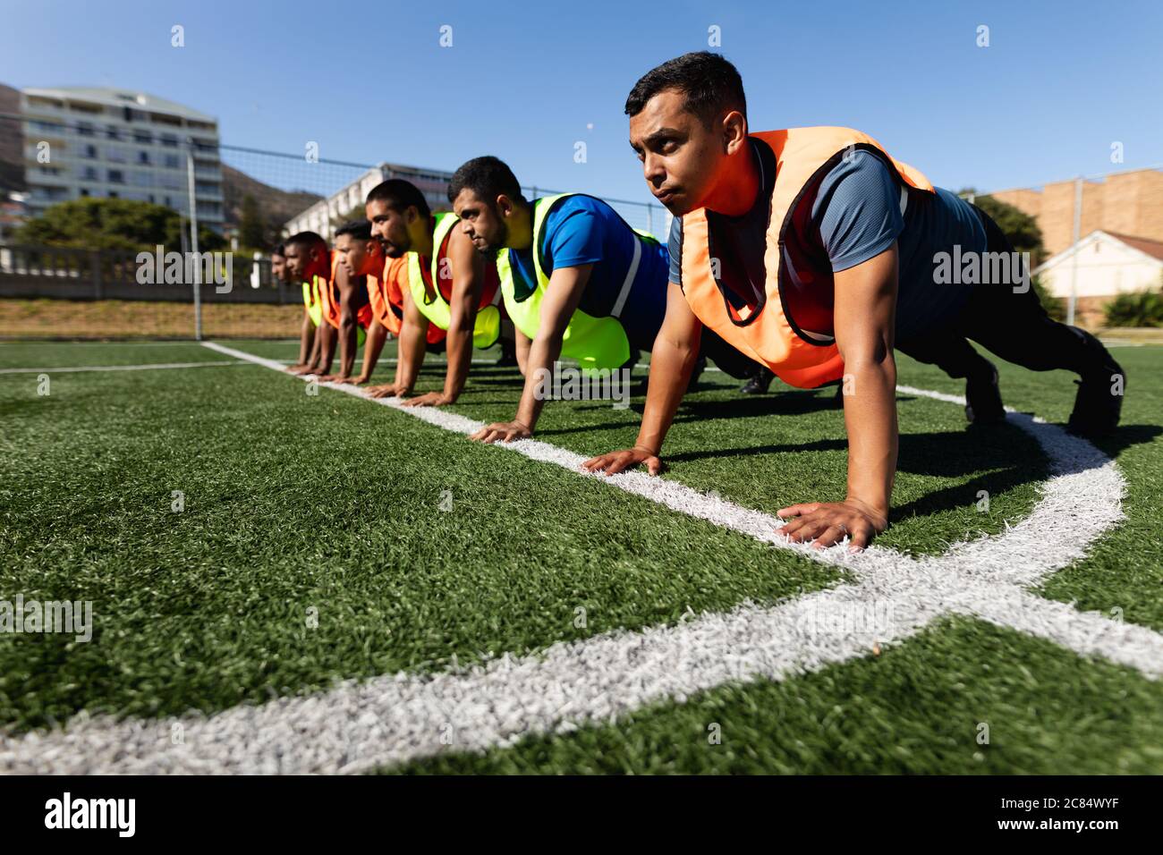 Multi ethnic group of male five a side football players wearing sports clothes and vests training at a sports field in the sun, warming up doing push Stock Photo