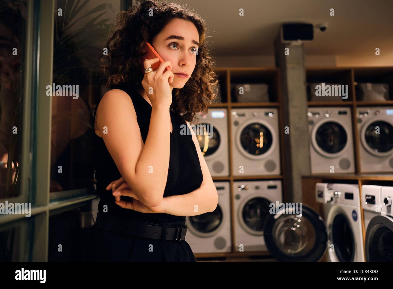 Beautiful girl talking on smartphone waiting washing in modern self-service laundry at night Stock Photo