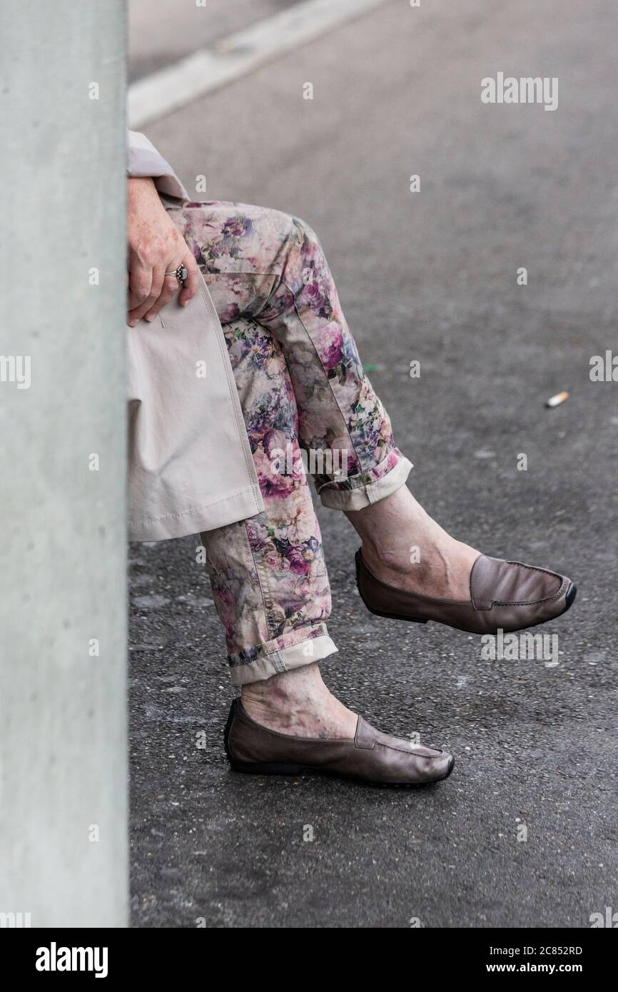 Woman sat on a railway station platform wearing a raincoat and patterned jeans with only her legs and hand showing beyond a concrete pillar Stock Photo