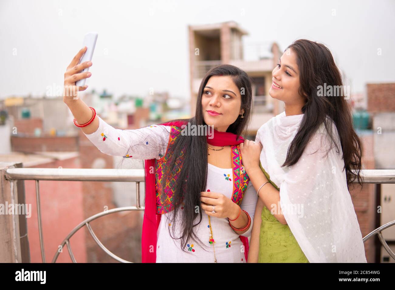 Beautiful Indian young women taking selfie together through smart phone in fresh air and spending their leisure at day time. Stock Photo