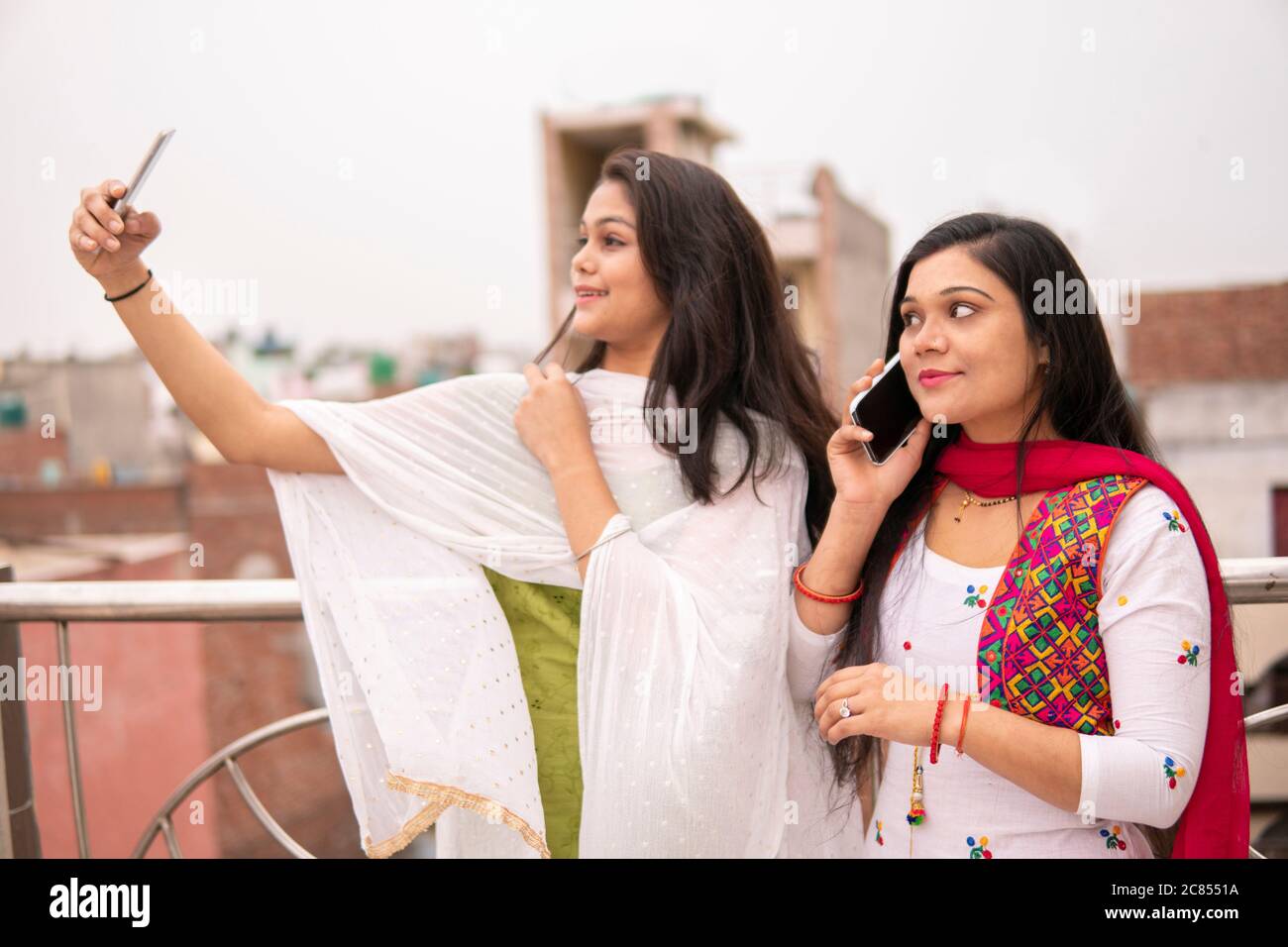Beautiful Indian young women standing together near railing. One woman is busy in taking selfie and other is talking on phone. Stock Photo