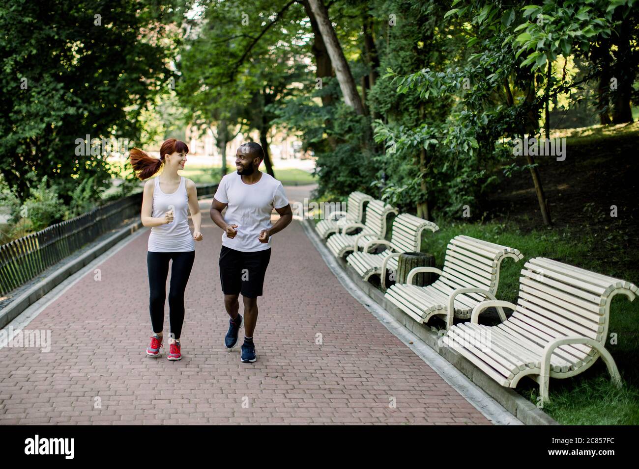 Happy joyful young multiethnic couple, African American man and Caucasian woman, jogging across the city park alley at early morning with sunrise and Stock Photo