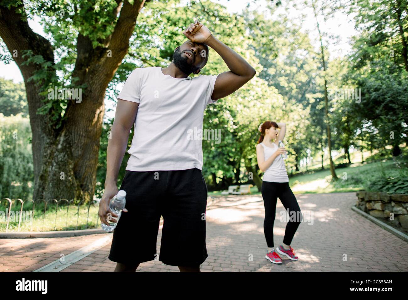 Sport lifestyle concept. Young strong African man tired after hard cardio workout, standing with water bottle. Exhausted Caucasian girl resting after Stock Photo