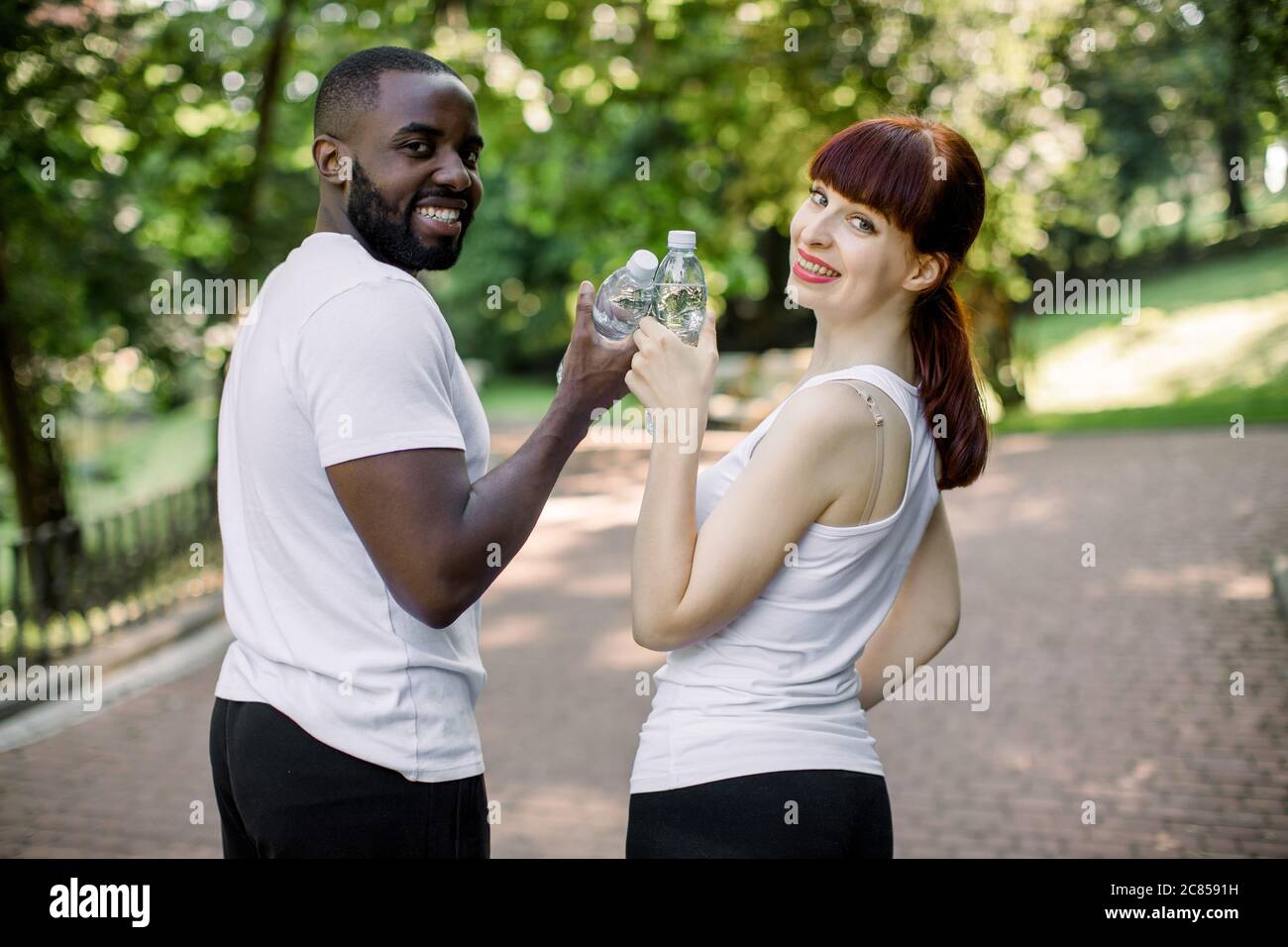 Sport, fitness, jogging and lifestyle concept. Cheerful smiling multiethnical couple with bottles of water, posing to camera during their break after Stock Photo