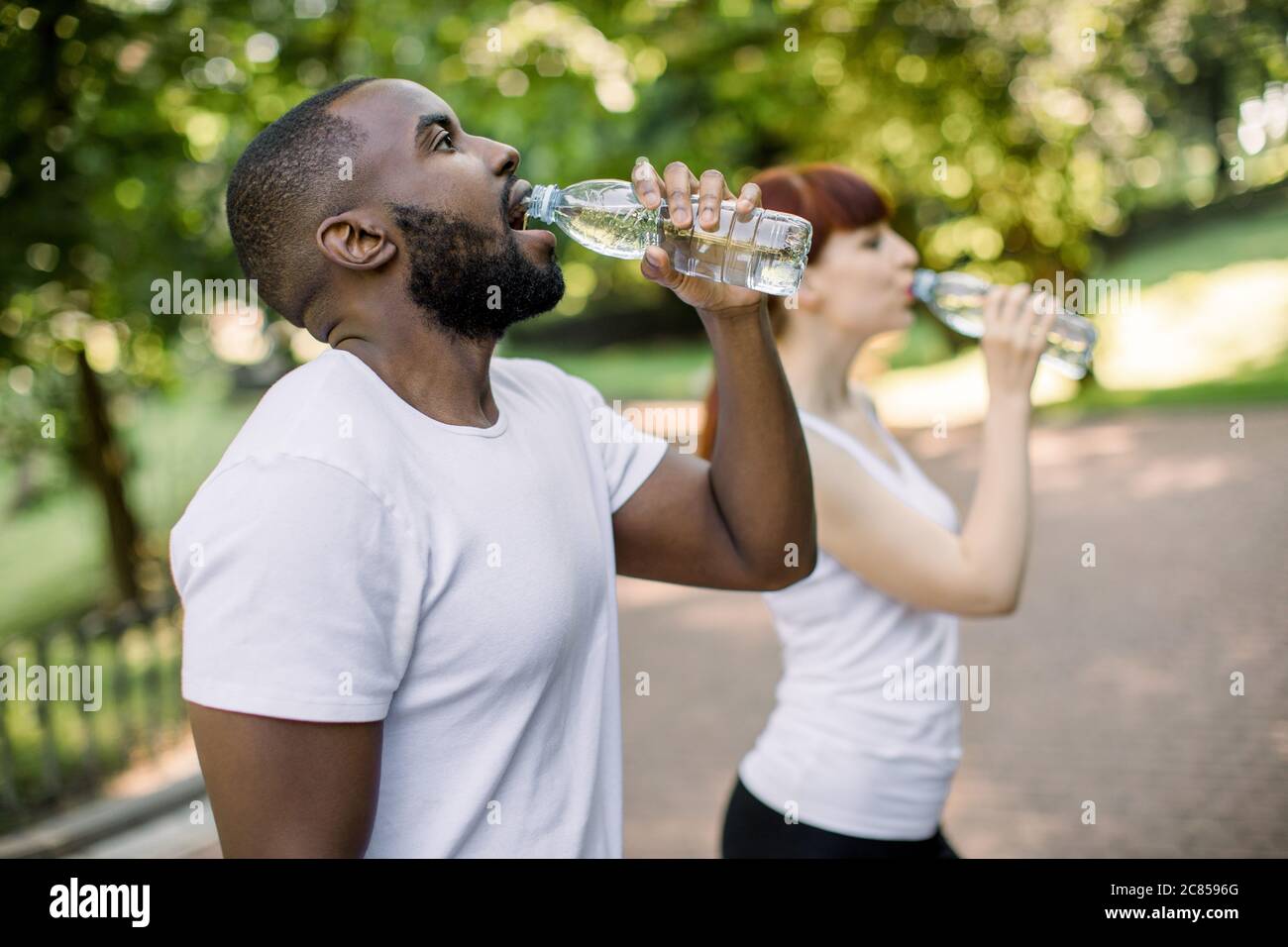 Multiethnical male and female friends drinking water from bottle after fitness sport exercise at city park. Smiling couple with bottles of cold drink Stock Photo