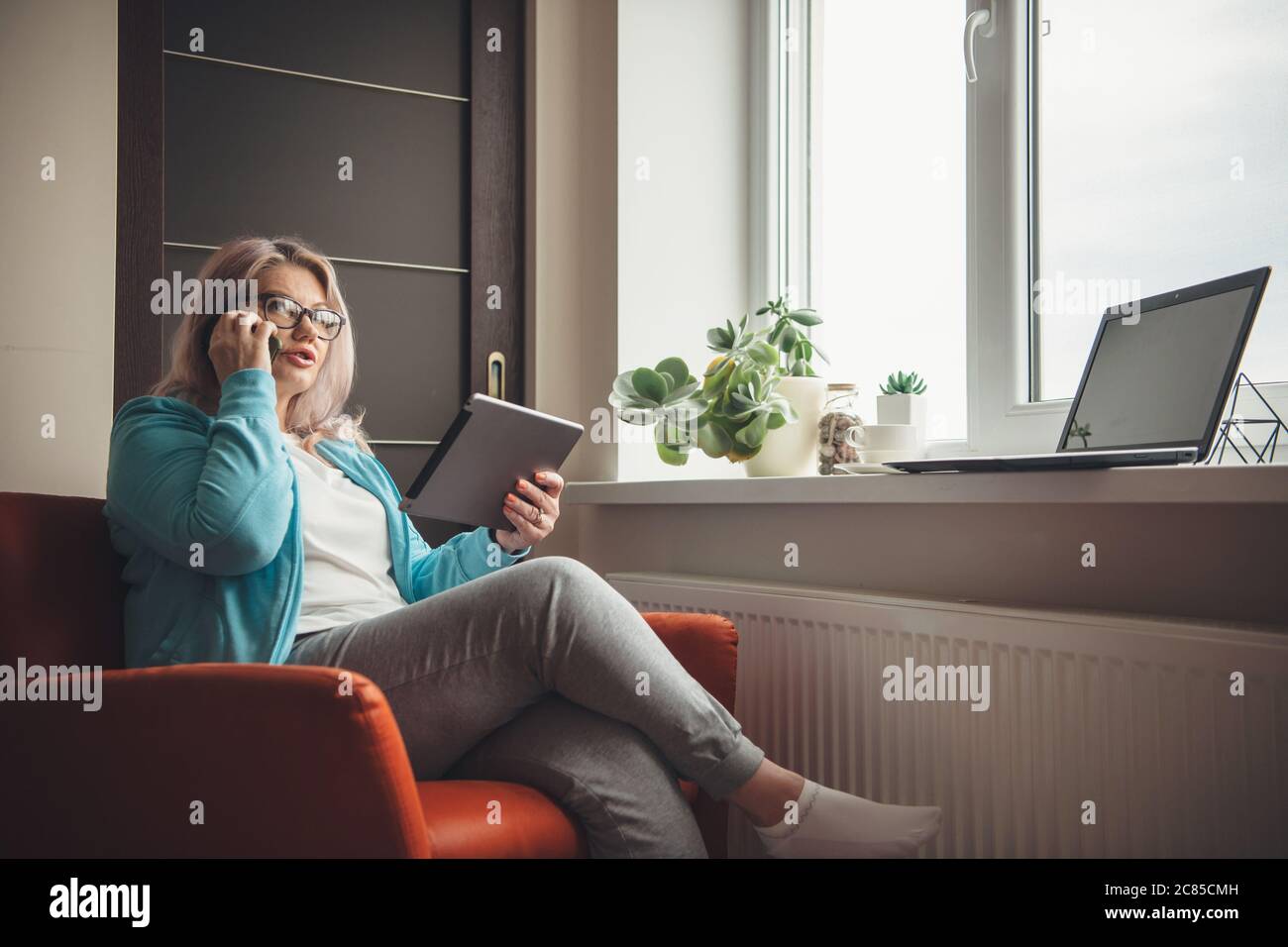 Busy senior woman with blonde hair and eyeglasses holding a tablet and talking on phone while working from home Stock Photo