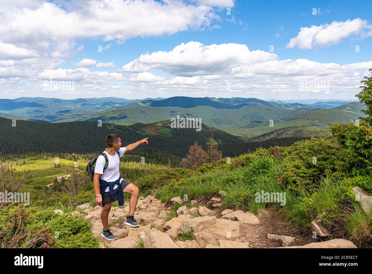 Man on peak of mountain. Emotional scene. Young man with backpack standing with raised hand on top of a mountain and pointing into the distance Stock Photo