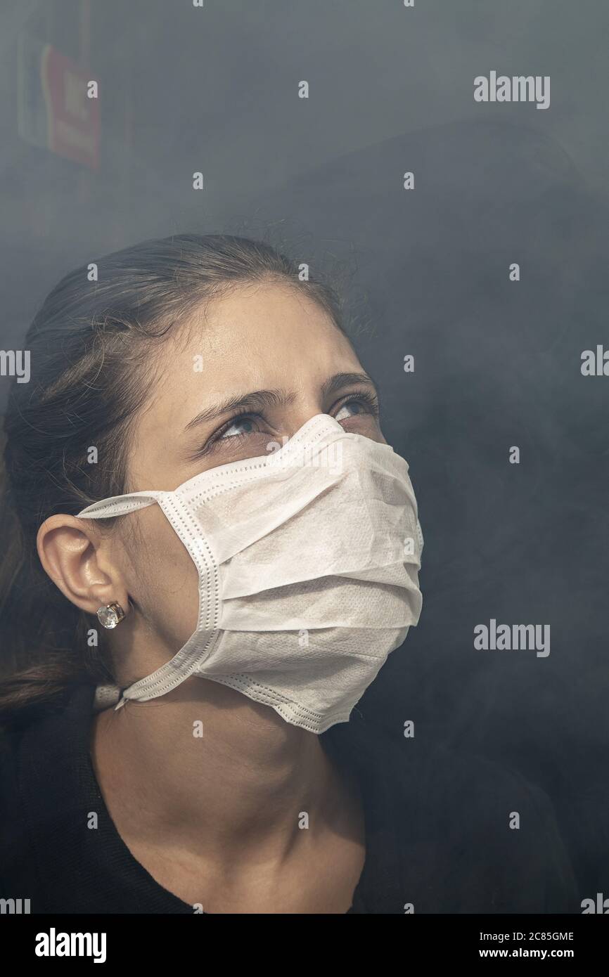 Vertical closeup shot of a Hispanic brunette female in a white medical mask surrounded by smoke Stock Photo