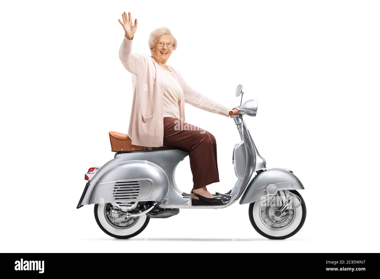 Profile shot of an elderly woman riding a vintage silver scooter and greeting with hand isolated on white background Stock Photo