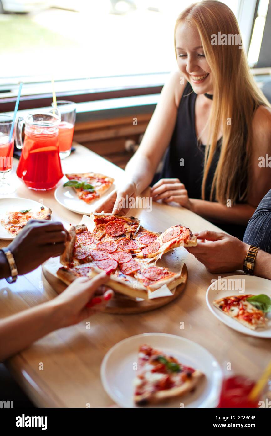 Summer dinner or lunch at street cafe indoor. Flat-lay of people hands taking freshly baked Italian pepperoni pizza with salami slices over white wood Stock Photo