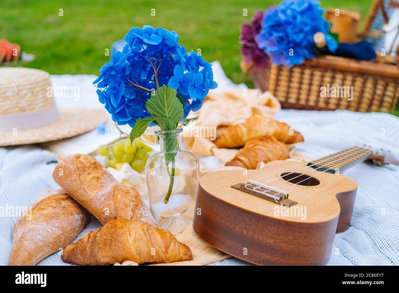 Summer picnic on sunny day with bread, fruit, bouquet hydrangea flowers,  glasses wine, straw hat, book and ukulele. Picnic basket on grass with food  a Stock Photo - Alamy