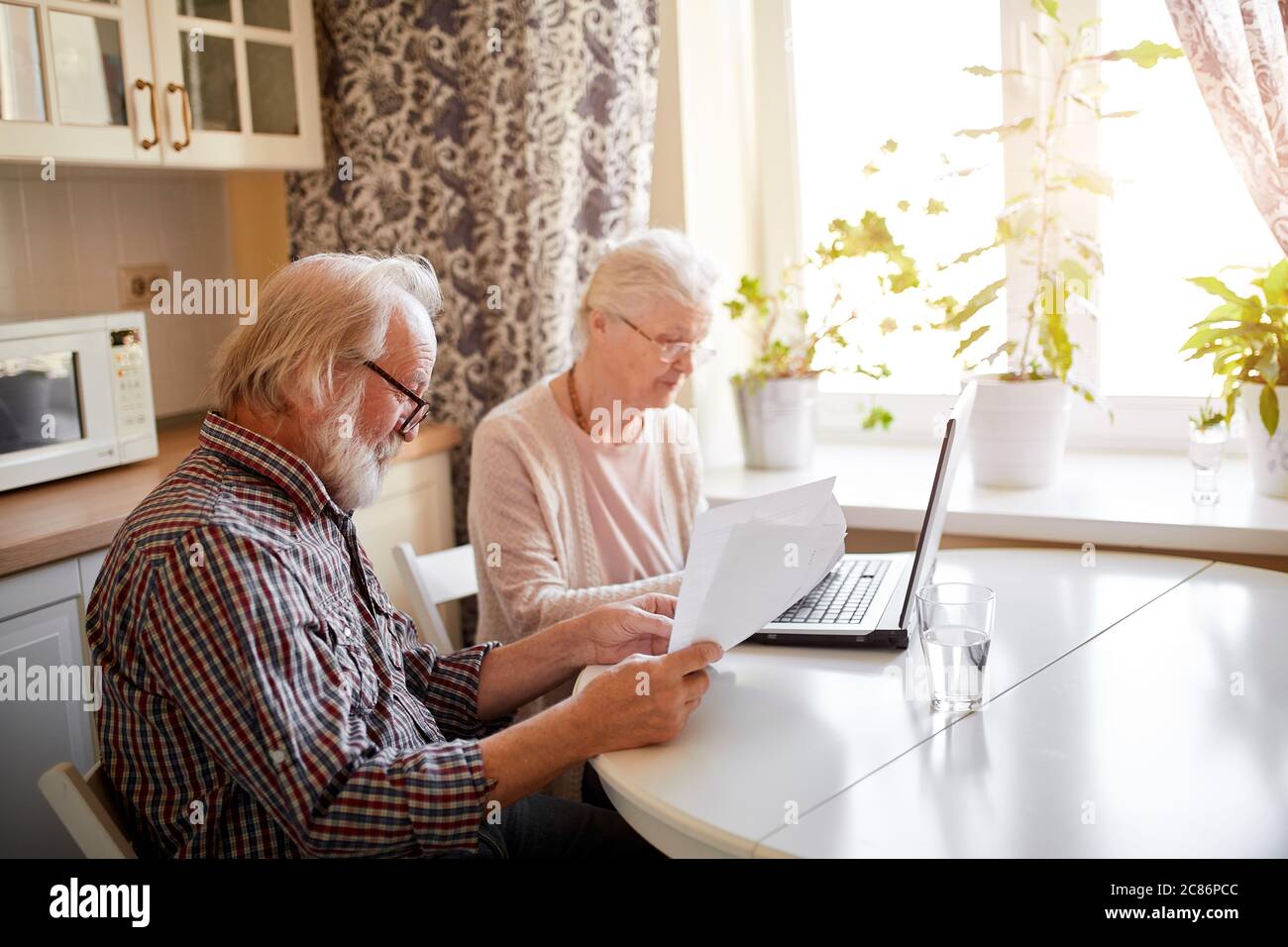 Elderly couple hold papers using laptop for online banking in cozy kitchen, satisfied senior couple smiling while browsing their family photos on digi Stock Photo