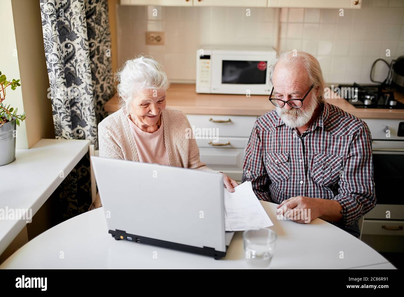 Elderly couple hold papers using laptop for online banking in cozy kitchen, satisfied senior couple smiling while browsing their family photos on digi Stock Photo