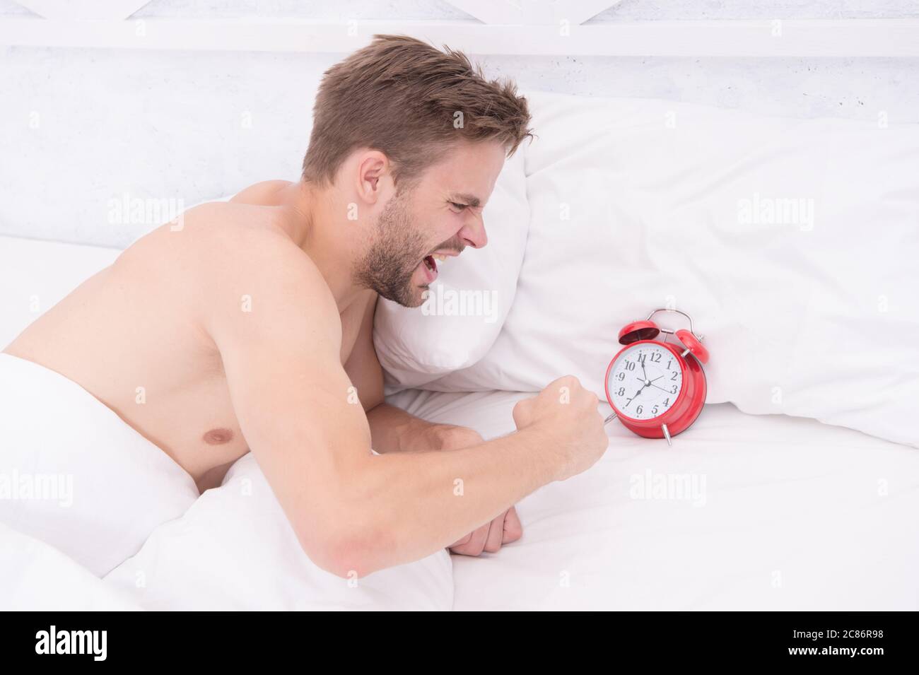 He is late. Angry guy break alarm clock in bed. Unshaven man awake from sleep. Keeping late hours. Wake timer. Wakeup time. Morning stress. Being late again. Stock Photo