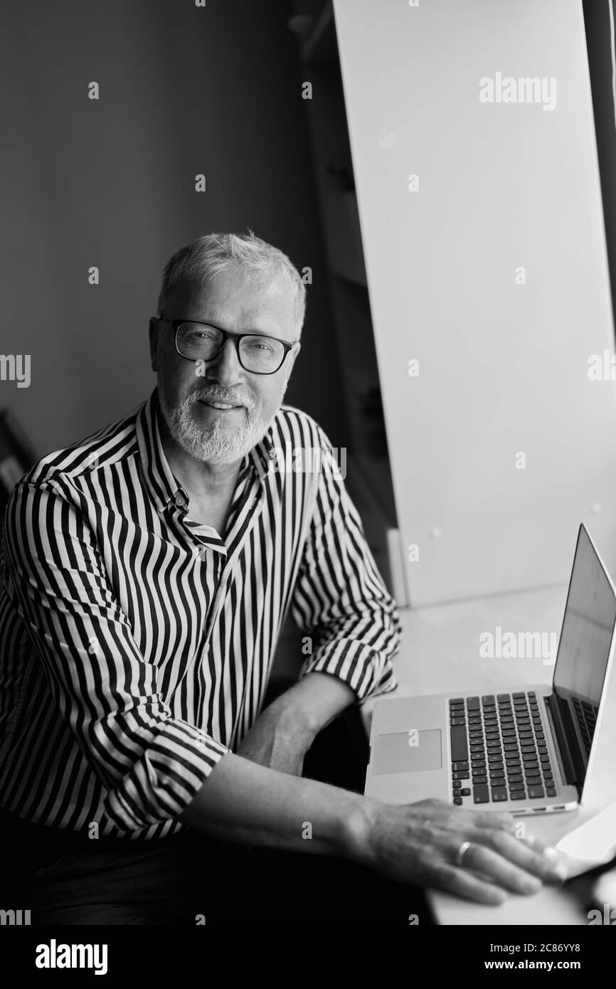 Trendy mature bearded man working from home with laptop. sitting at desk near window Stock Photo