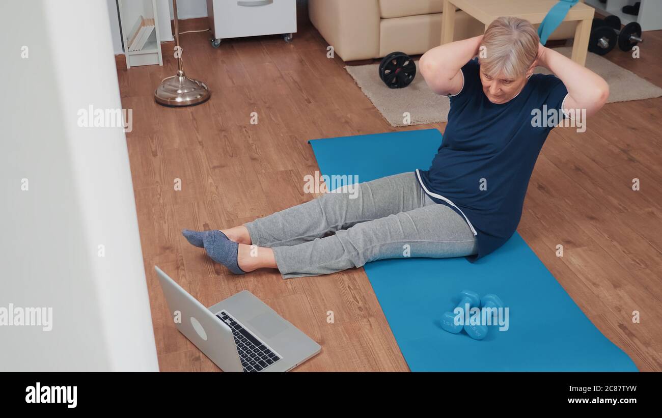 Sportive senior woman on yoga mat exercising looking at online fitness program. Old person pensioner online internet exercise training at home sport activity at elderly retirement age Stock Photo
