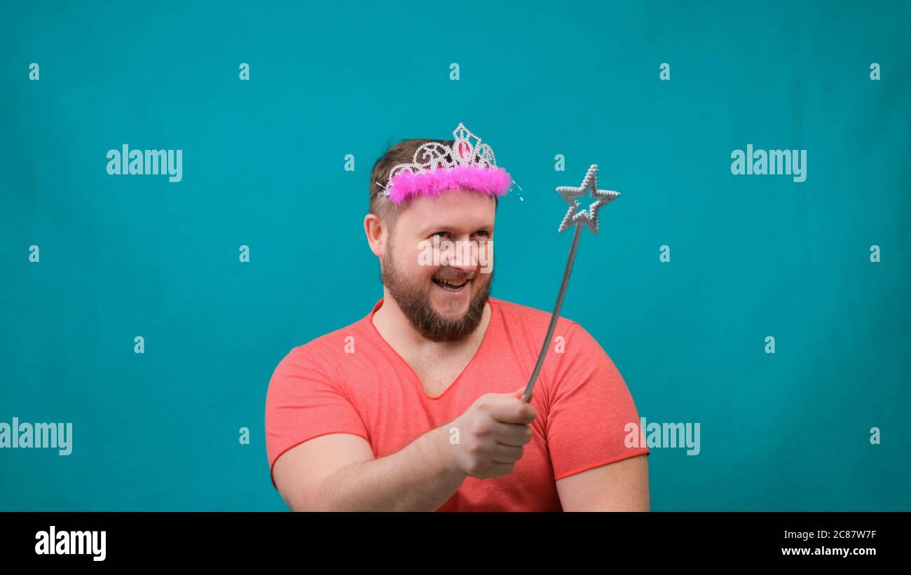 funny bearded freaky man in a pink T-shirt with a deadema on his head is dreaming with a magic wand in his hand. A funny wizard joke to make and Stock Photo