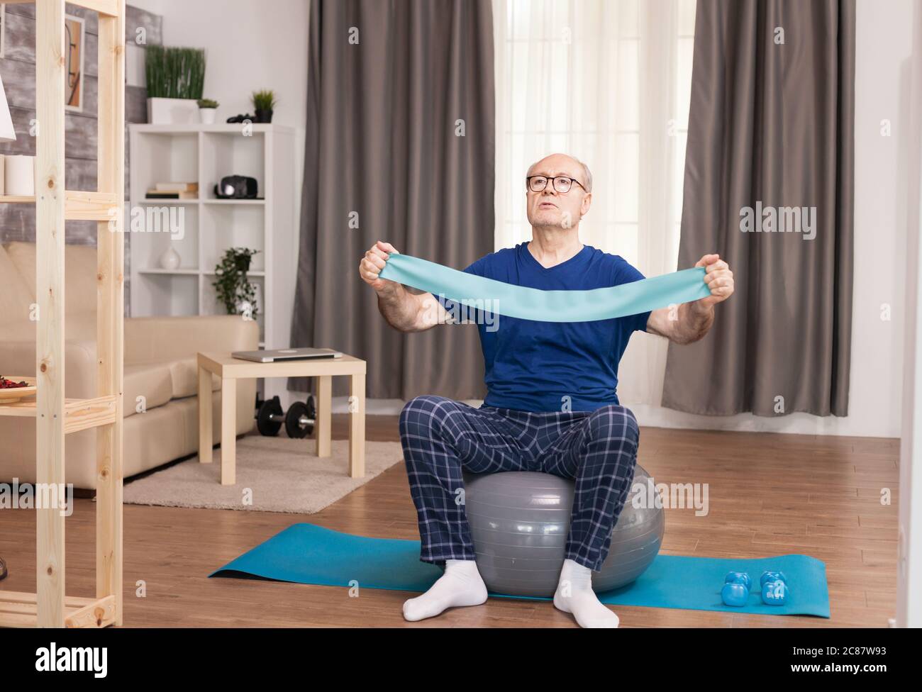 Old man using the resistance band during his training. Old person pensioner online internet exercise training at home sport activity with dumbbell, resistance band, swiss ball at elderly retirement age. Stock Photo