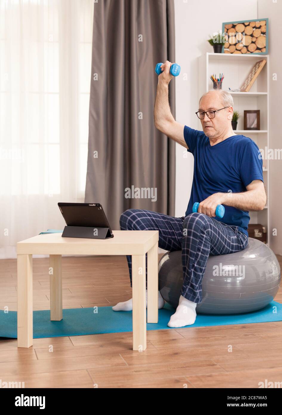 Senior man doing muscle recovery exercises sitting on a swiss ball in front of the tablet. Old person pensioner online internet exercise training at home sport activity with dumbbell resistance band s...