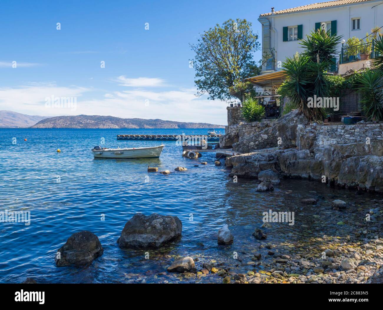 Pebble sea shore and The White House in Kalami, former residence of British authors Lawrence and Gerald Durrell and their family. Boat and pier at Stock Photo