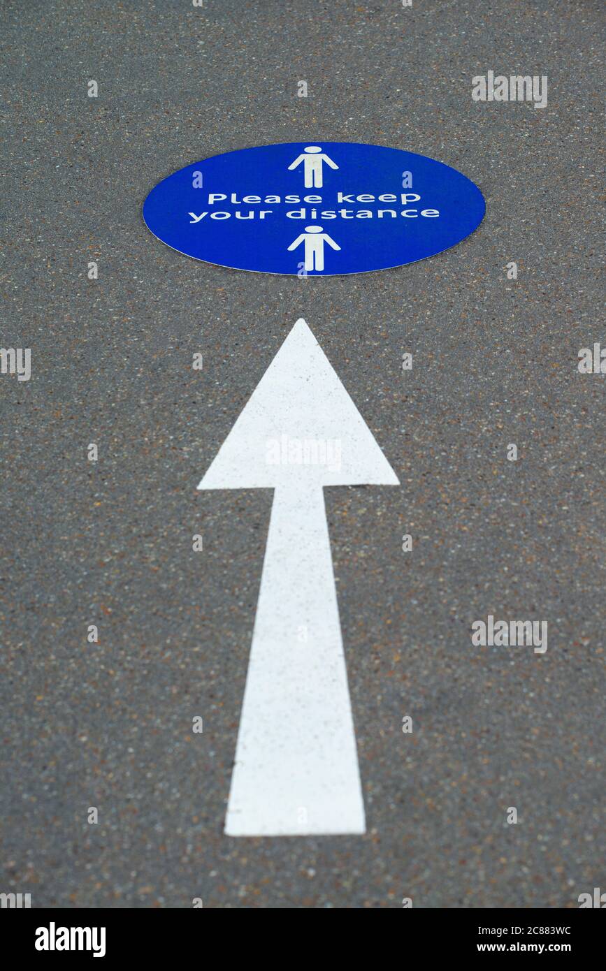 White directional arrow symbol with blue notice on pavement, advising pedestrians to keep to the social distancing rules. UK Coronavirus pandemic sign. Stock Photo