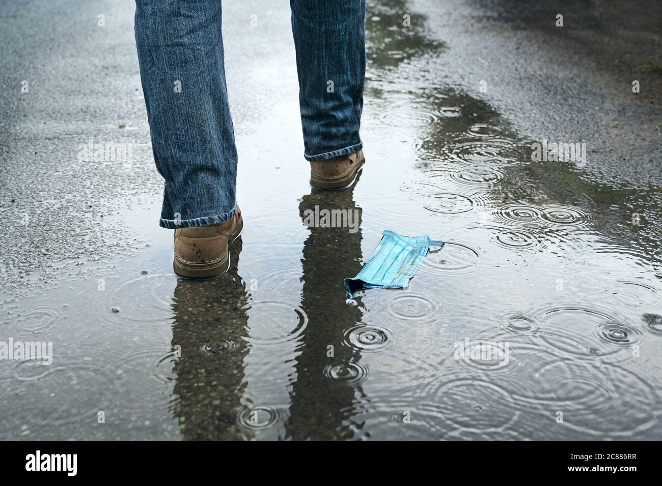 Medical face mask discarded on the asphalt in the rain, man walks away, thoughtless against the danger of coronavirus infection after the lockdown, co Stock Photo