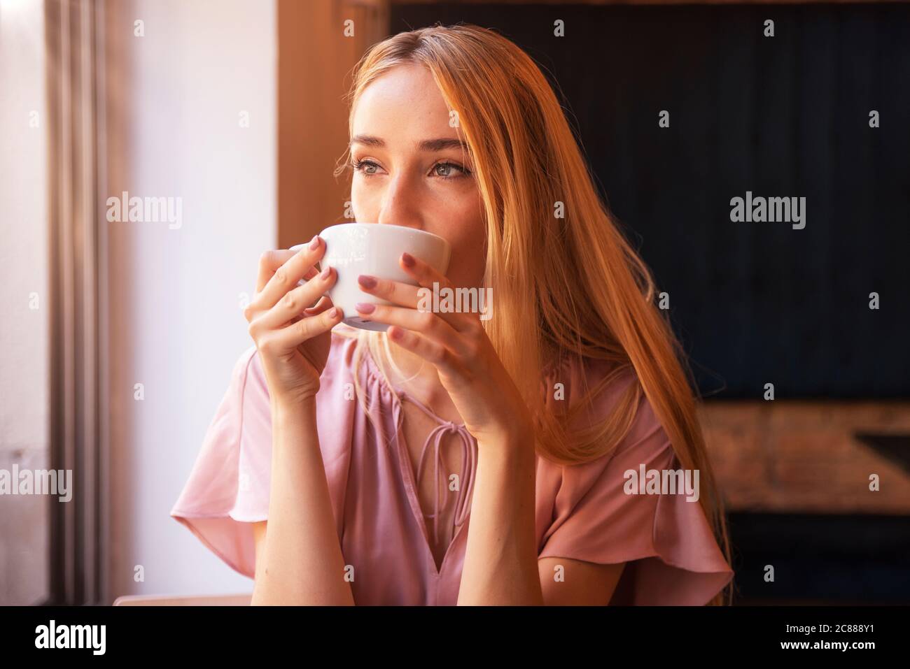 Portrait of young woman sitting at cafe with cup of coffee and looking away while daydreaming. Stock Photo