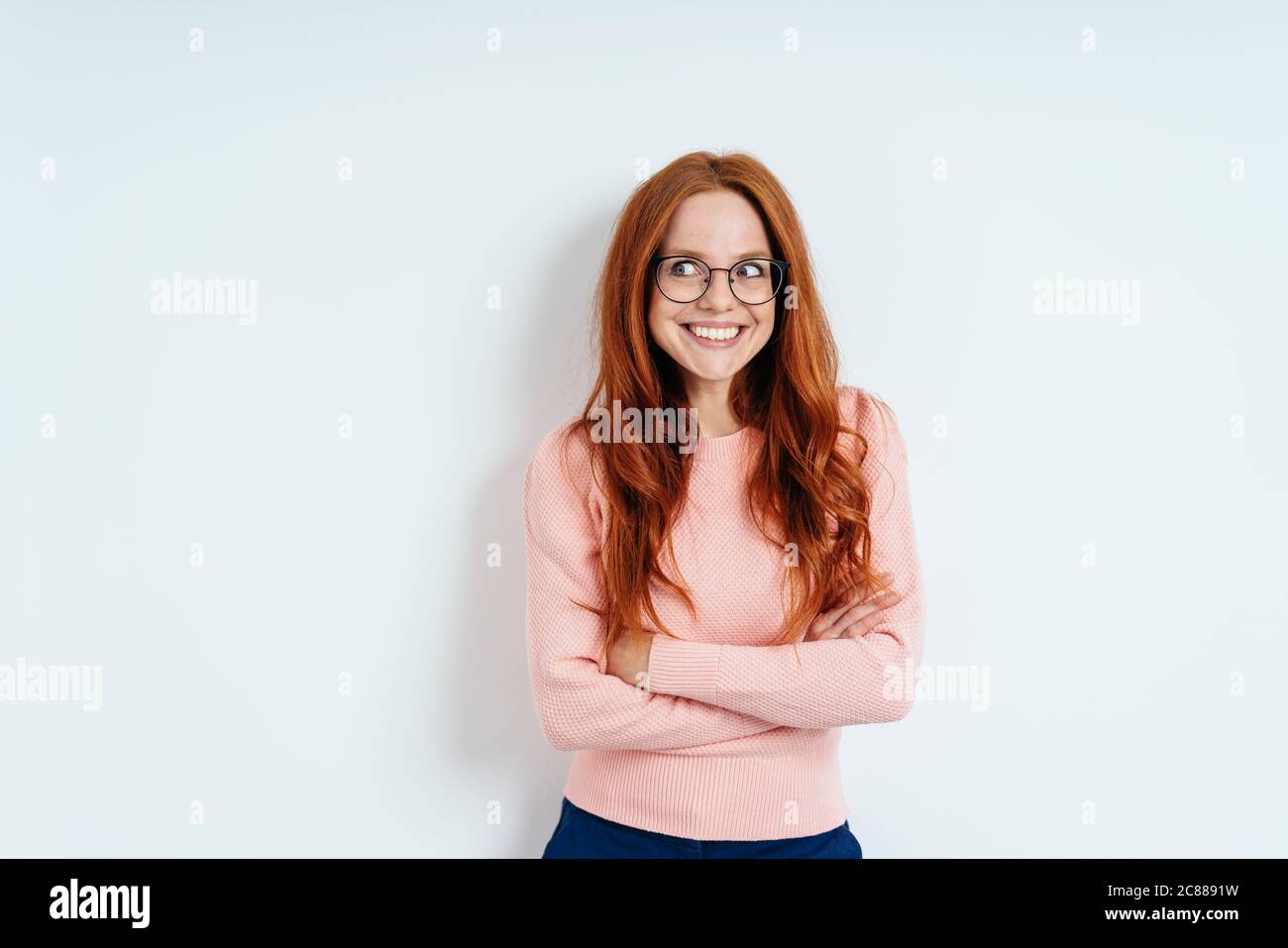 Gleeful young woman looking aside in anticipation with an eager expression and folded arms against a white interior wall with copy space Stock Photo