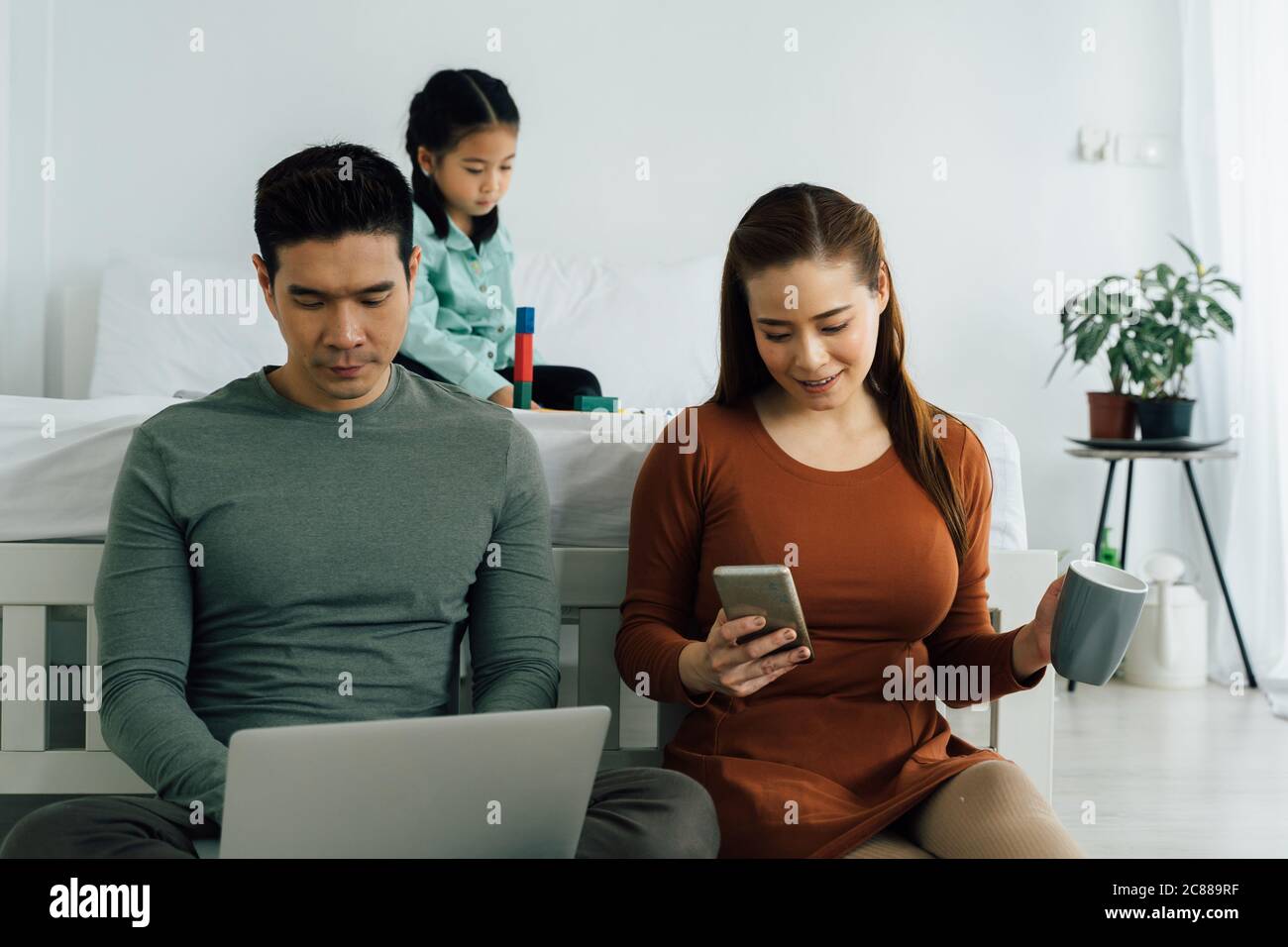 Cheerful couple sitting with computer and smartphone as young girl plays alone in the background Stock Photo