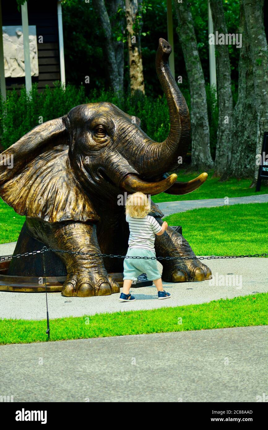 Matakana, New Zealand - Dec 2019. Bronze sculpture representing elephant climbing out of a manhole, and cute little child fascinated by the elephant Stock Photo