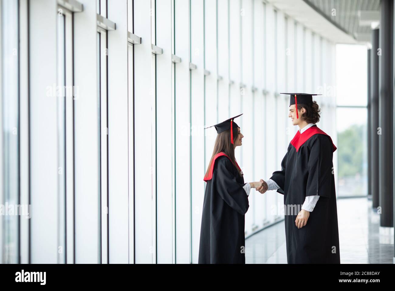 Happy international students in mortar boards and bachelor gowns greeting each other by handshake in University Stock Photo