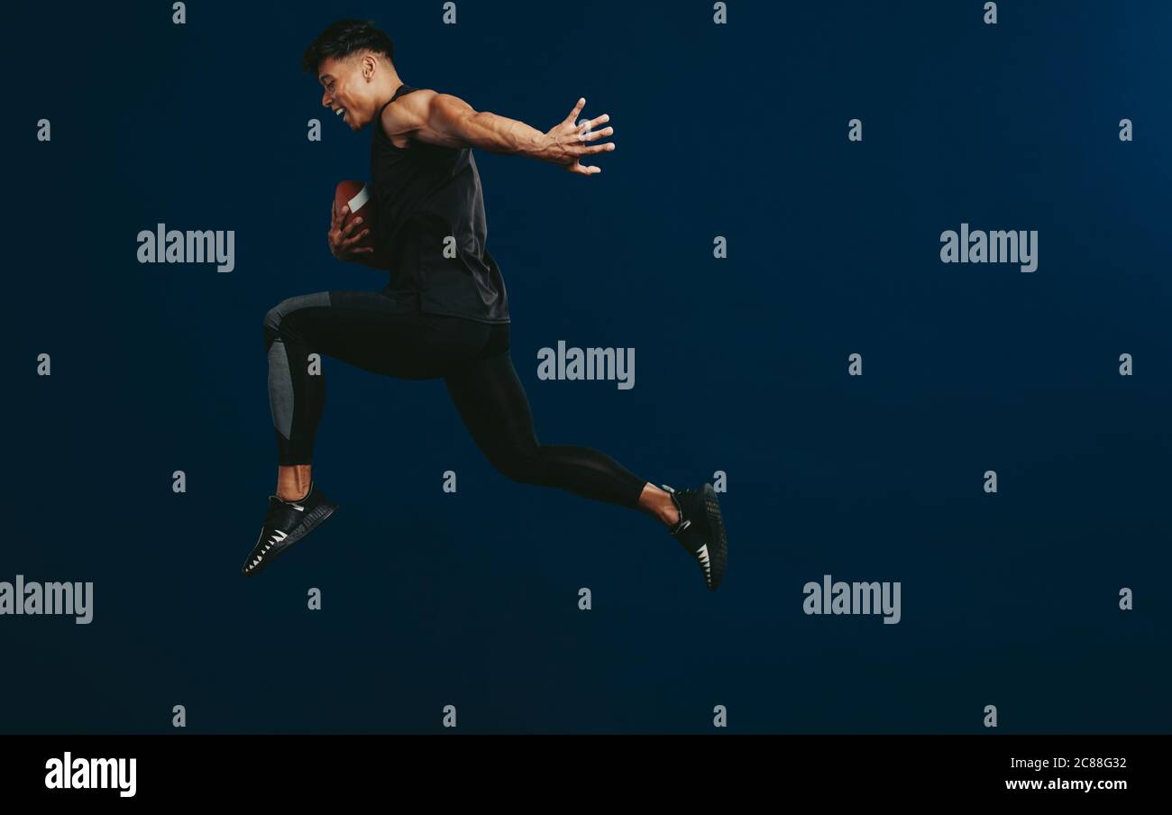 Smiling American football player holding ball and jumping against dark background. Sportsman forward to the victory with a football in hand. Stock Photo
