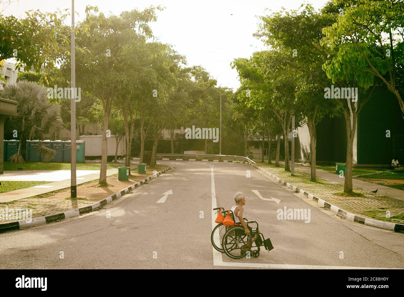 A chinese old man in singlet pushing his own wheelchair to cross the road. Stock Photo