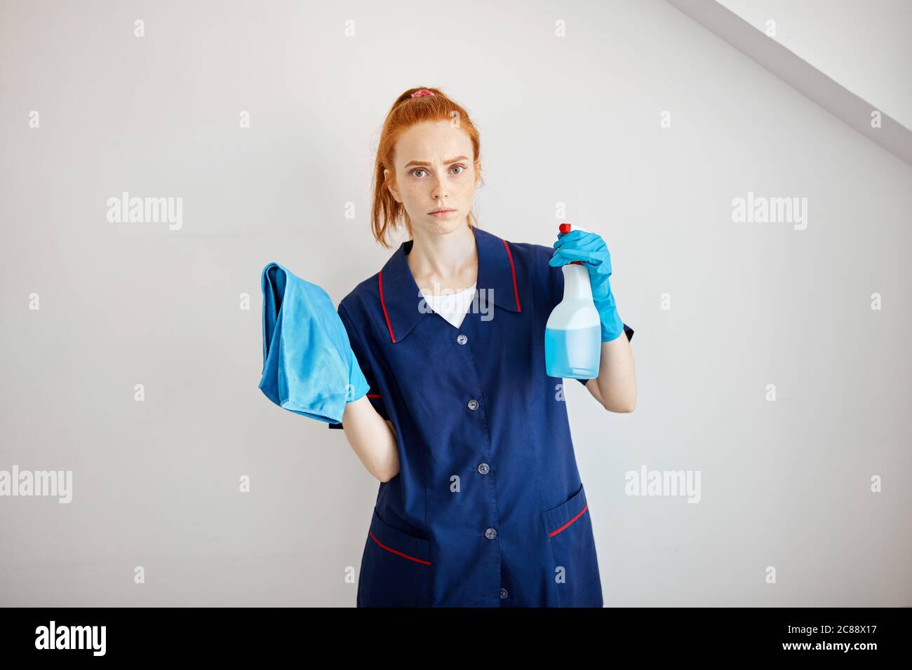 Beautiful red-haired housewife wearing household rubber gloves and blue dressing gown looking at camera posing with blue dusting cloth and wiper. Home Stock Photo