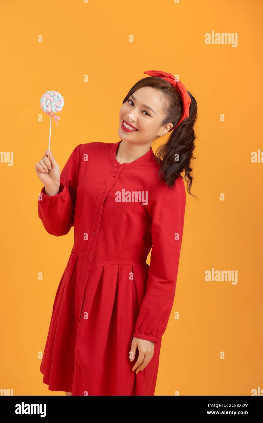 Smiling young Asian woman in red dress holding lollipop while standing over orange backgound. Stock Photo