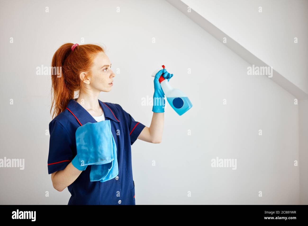 Beautiful red-haired housewife wearing household rubber gloves and blue dressing gown looking at camera posing with blue dusting cloth and wiper. Home Stock Photo
