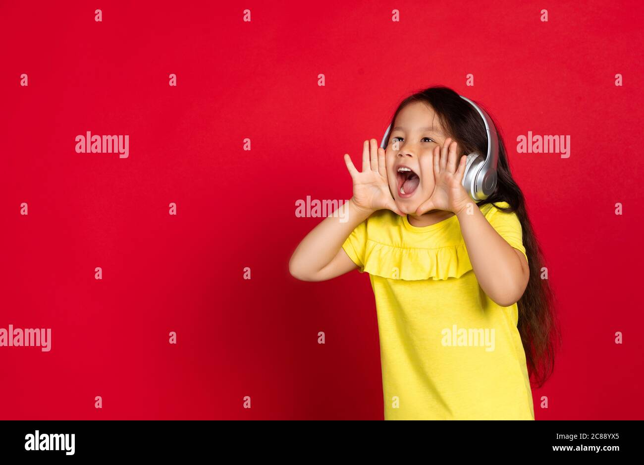 Shouting for sales, wearing headphones. Beautiful little girl on red background. Half-lenght portrait of happy child. Cute asian girl in yellow. Concept of facial expression, human emotions, childhood. Stock Photo