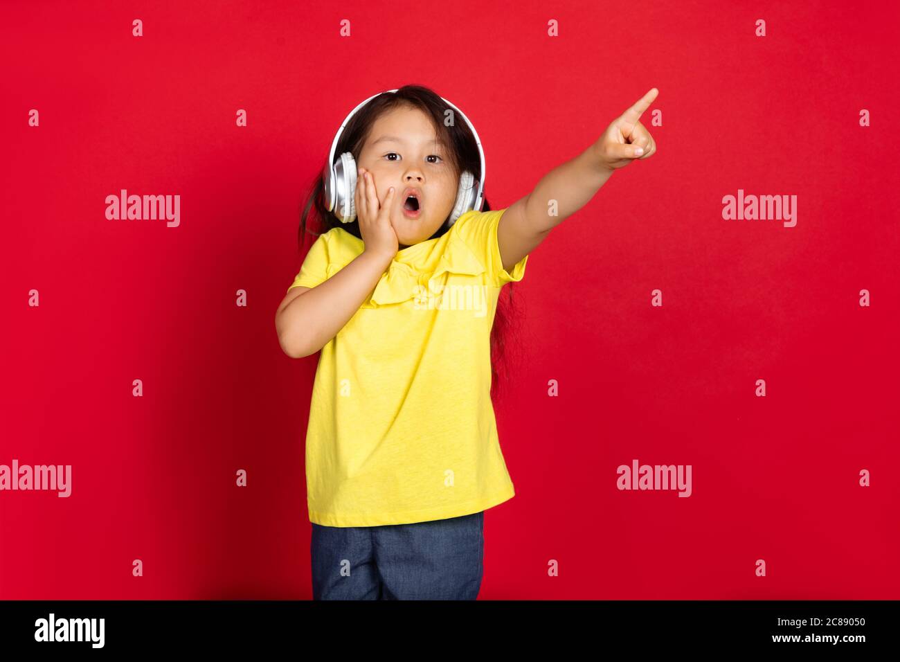 Pointing shocked, wearing headphones. Beautiful little girl on red background. Half-lenght portrait of happy child. Cute asian girl in yellow. Concept of facial expression, human emotions, childhood. Stock Photo