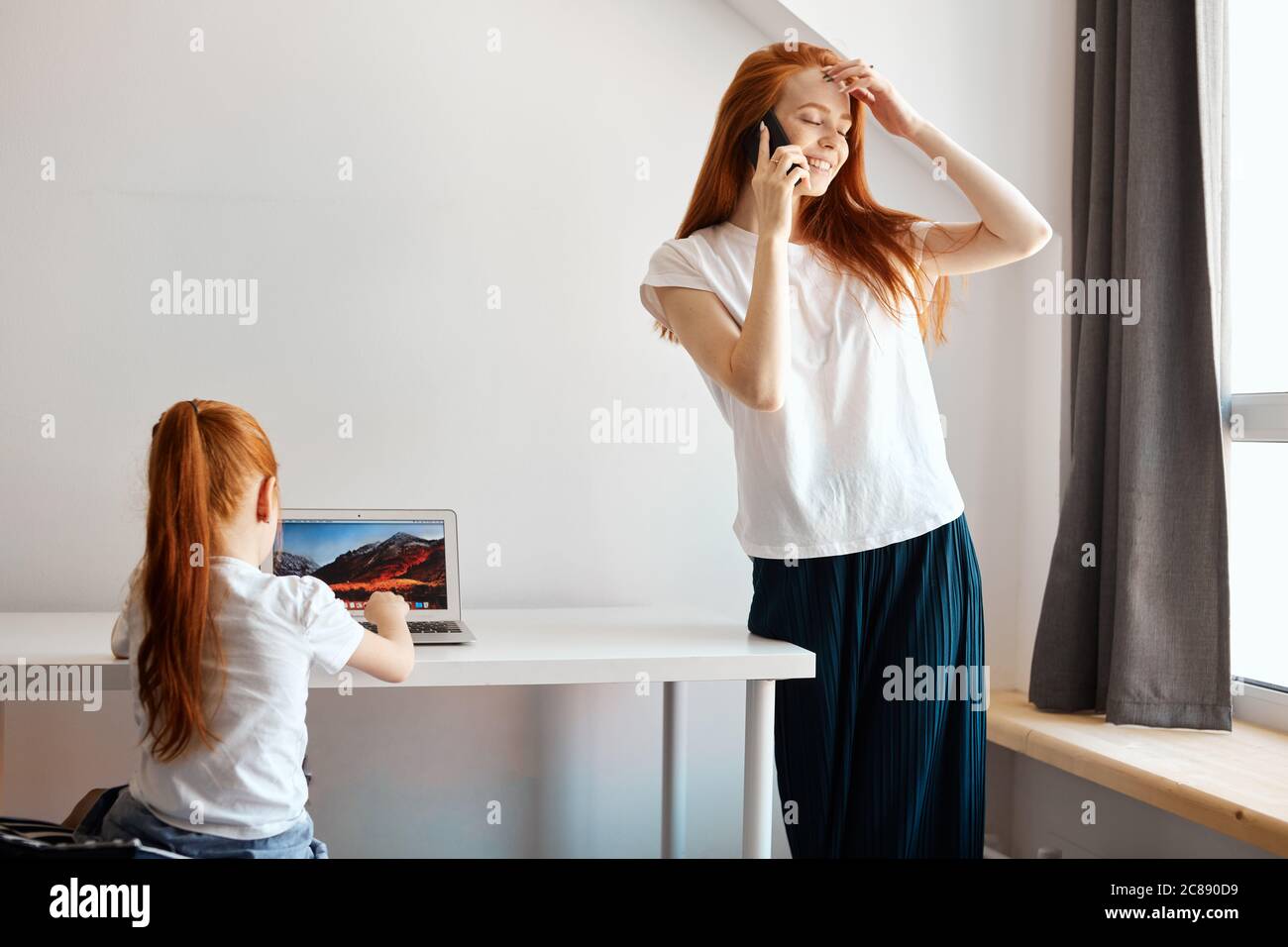 Red-haired preschool age daughter sitting at table and looking at the laptop, watching new TV chanal for kids while her content Mom smiling and talkin Stock Photo