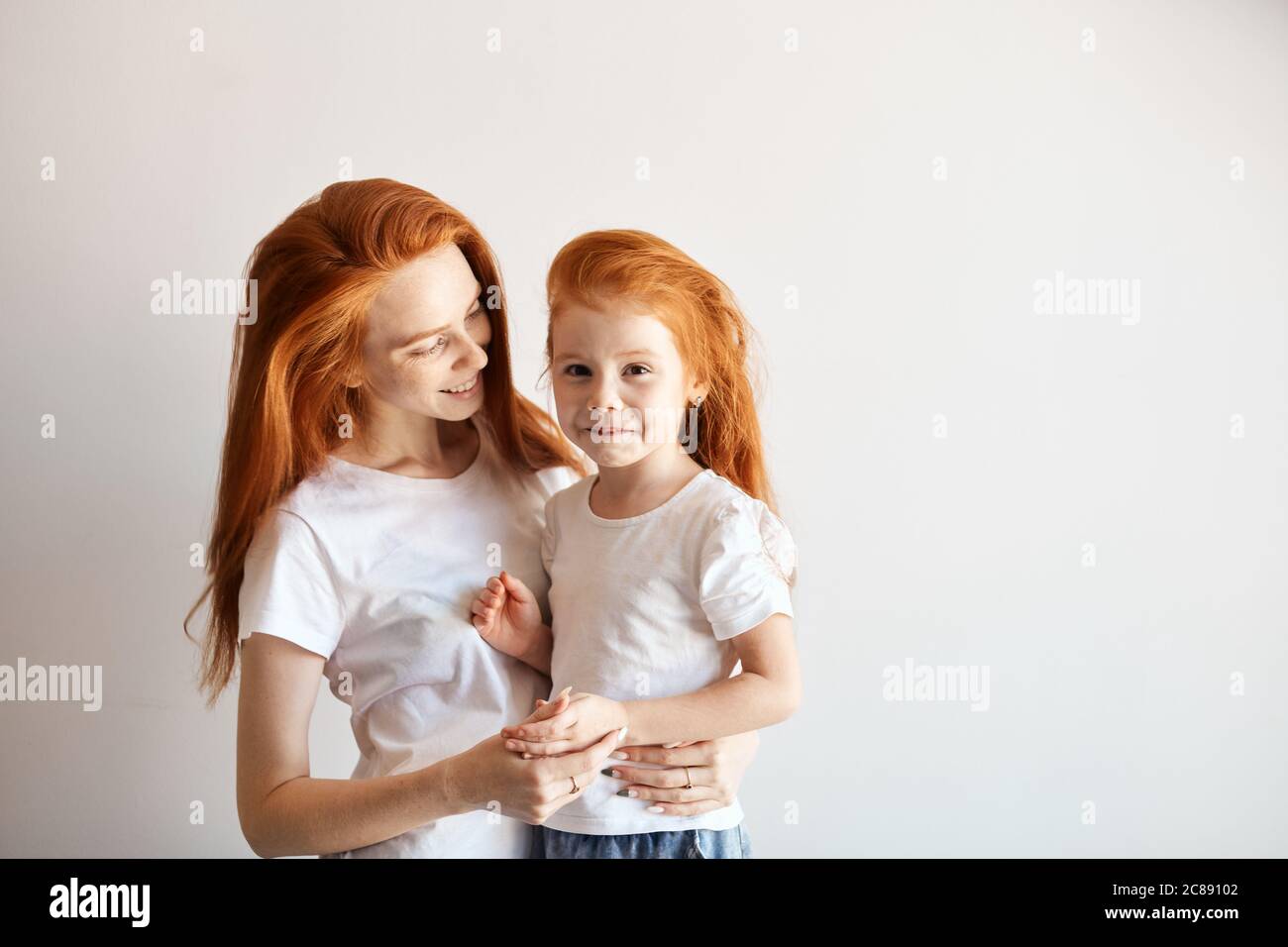 Close-up portrait of smiling mother hugging cute four year old daughter isolated on white background. Two red headed similar females of different gene Stock Photo