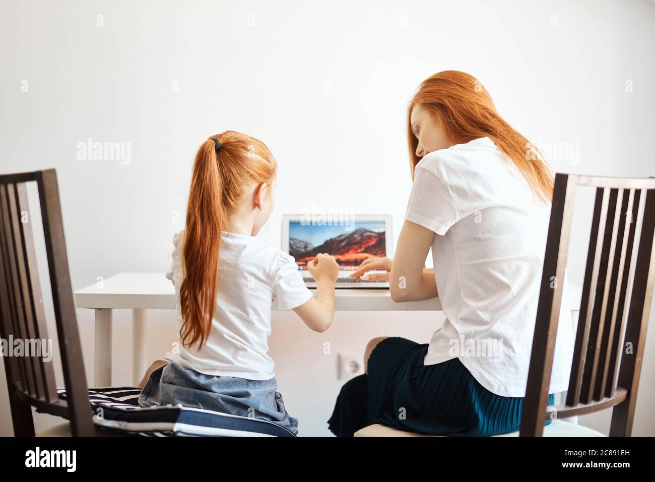 Loving caucasian red-haired mummy and her, sweetheart girl sitting at table and looking at the laptop, watching new TV chanal for kids, back view. Stock Photo