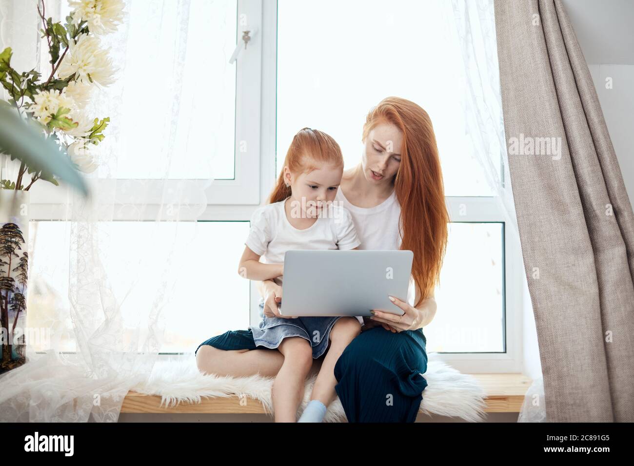 Loving caucasian red-haired mummy and her, sweetheart girl sitting at table and looking at the laptop, watching new TV chanal for kids sitting on wind Stock Photo