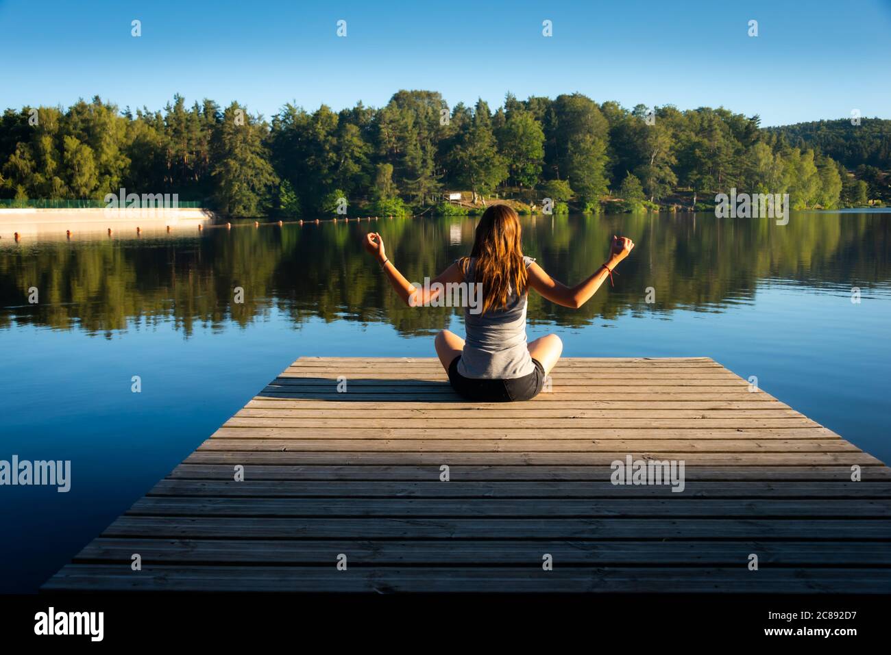 young woman on wooden pontoon or pier practicing yoga & relaxing on holiday . Stock Photo