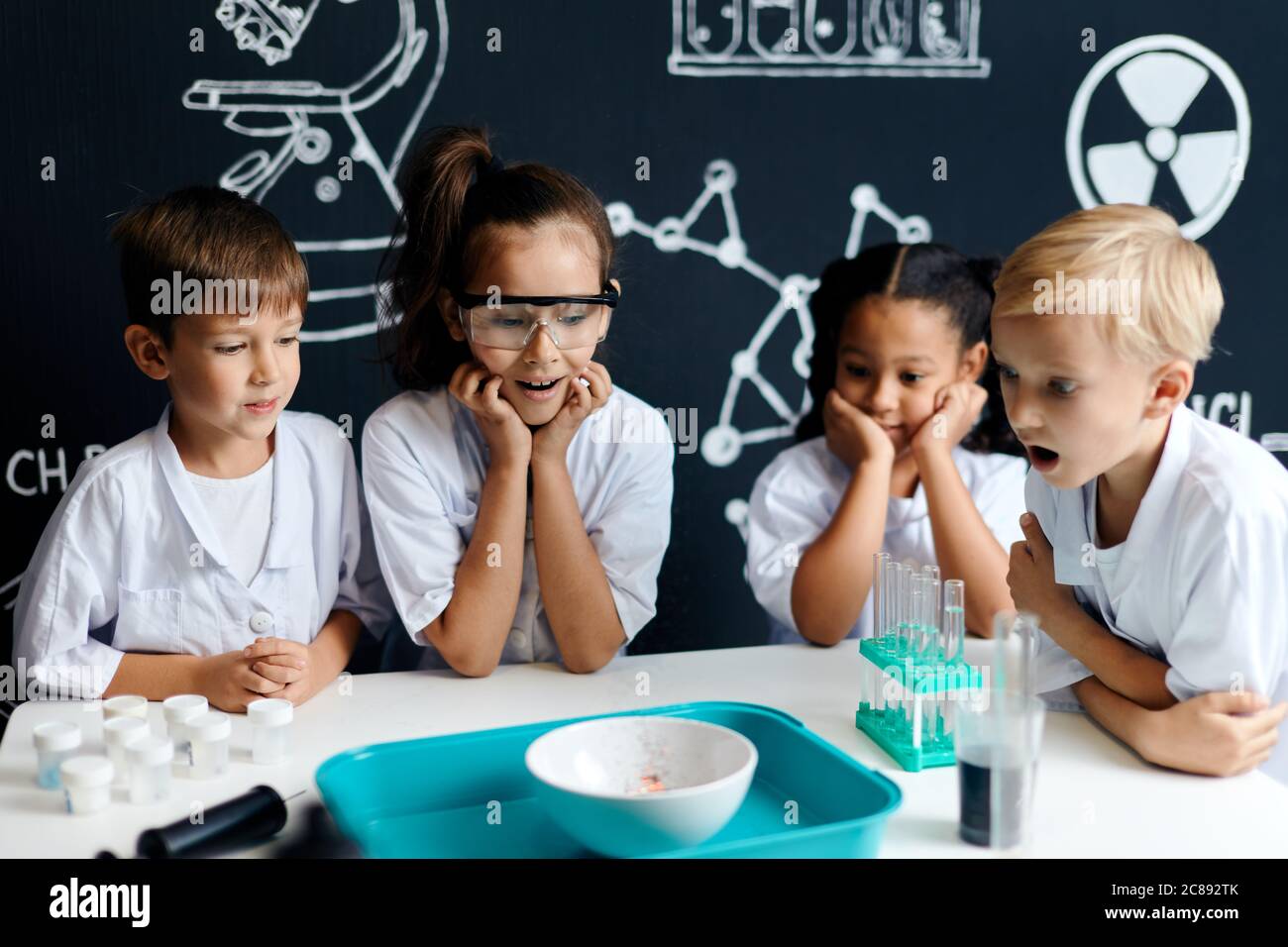 Miracles of chemistry. A group of cheerful pre-teen pupils in lab coats gathering near the table and observing the chemical reaction going on in the b Stock Photo