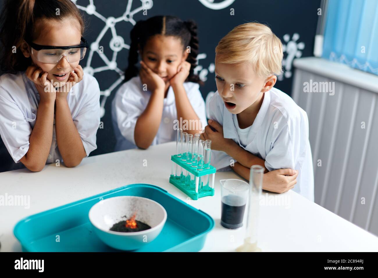 Group of multiracial diverse kids in scientist white clothes doing science experiments with reagents in the laboratory, being surprised and astonished Stock Photo
